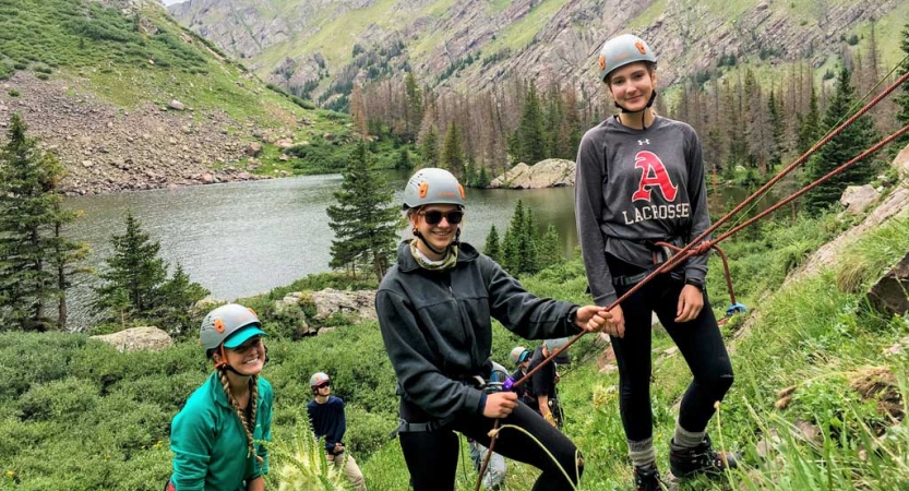 Three people wearing safety gear stand in a grassy area and smile for the photo. one is attached to ropes and appears to be belaying a climber who is not pictured. 